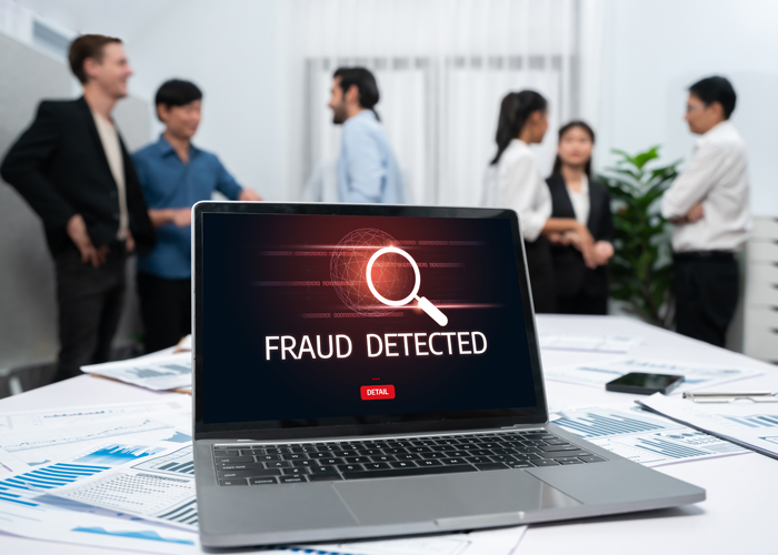 The screen on the laptop shows 'Fraud detected', people talking behind the laptop